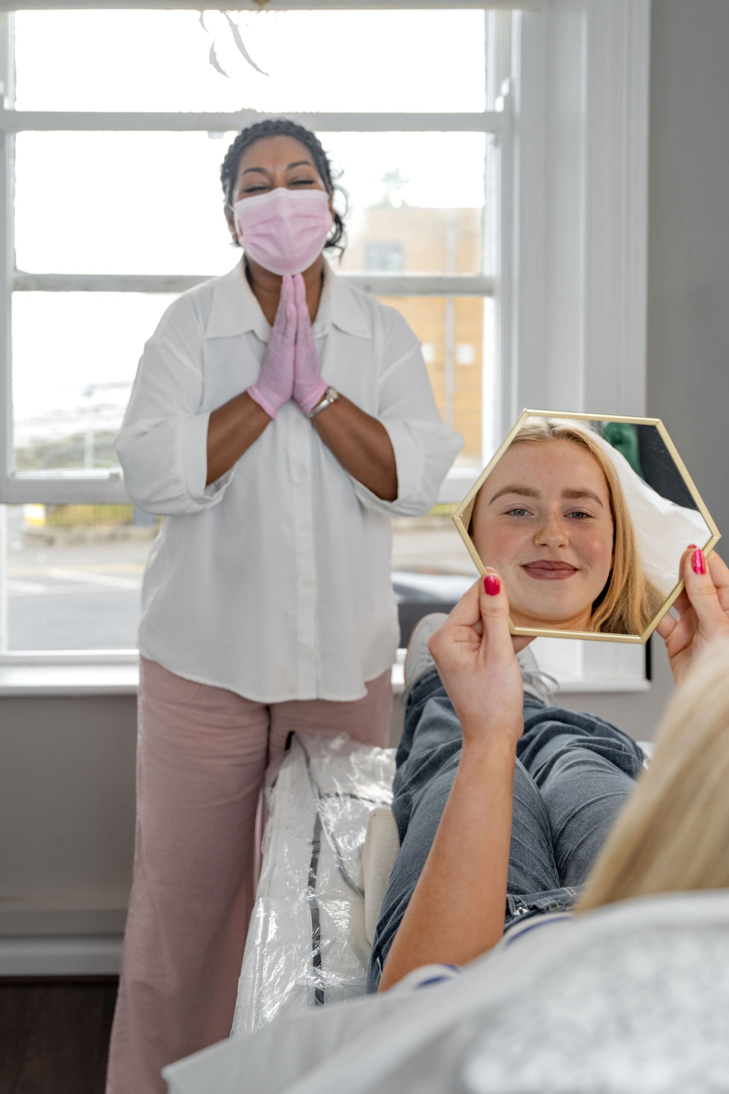 A brow treatment office scene with a smiling woman in a white coat and pink face mask standing behind a seated patient who is holding a hexagonal mirror to check her eyebrows. The patient has pink nail polish, and the background shows a bright window.