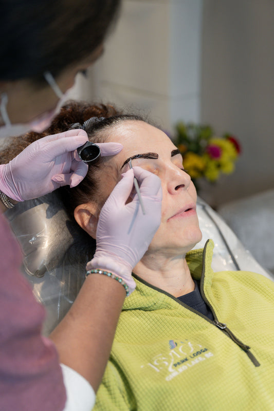 A close-up lifestyle shot of a brow lamination treatment in progress. A client lies in a spa chair while a technician, wearing pink gloves and a face mask, carefully processes the client's eyebrow.