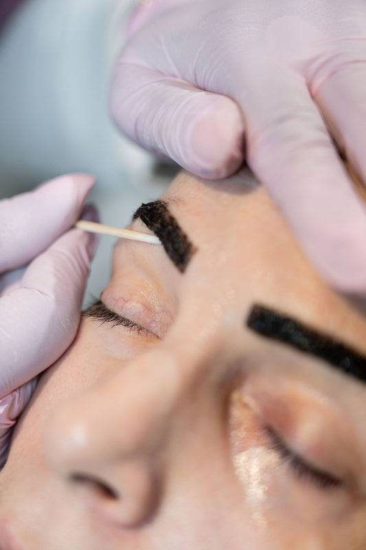 Close-up of a person's face with closed eyes while another person wearing pink gloves applies dark eyebrow makeup or tattoo pigment with a small wooden tool.