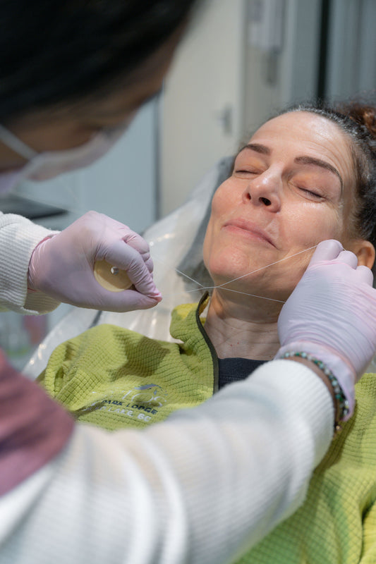 A close-up lifestyle shot of a woman receiving a chin thread treatment, lying down with her eyes closed while a practitioner in white gloves and a face mask applies the thread to her chin.