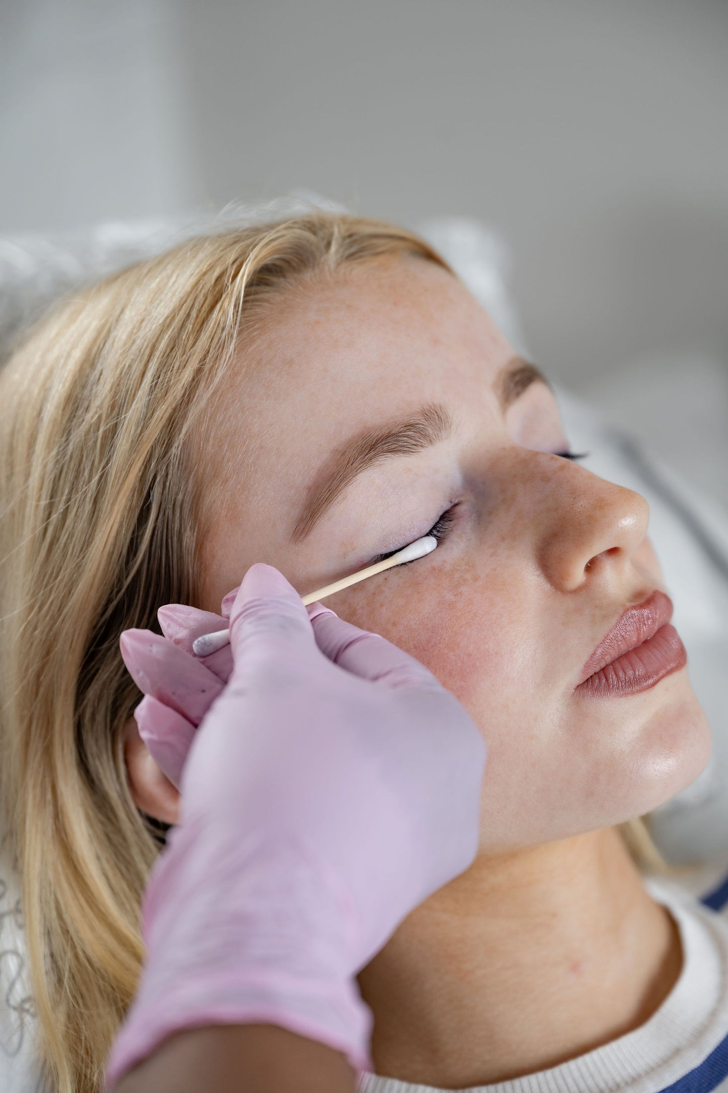 A close-up lifestyle shot of a beauty therapist wearing a pink glove using a cotton swab on a blonde woman's eye.