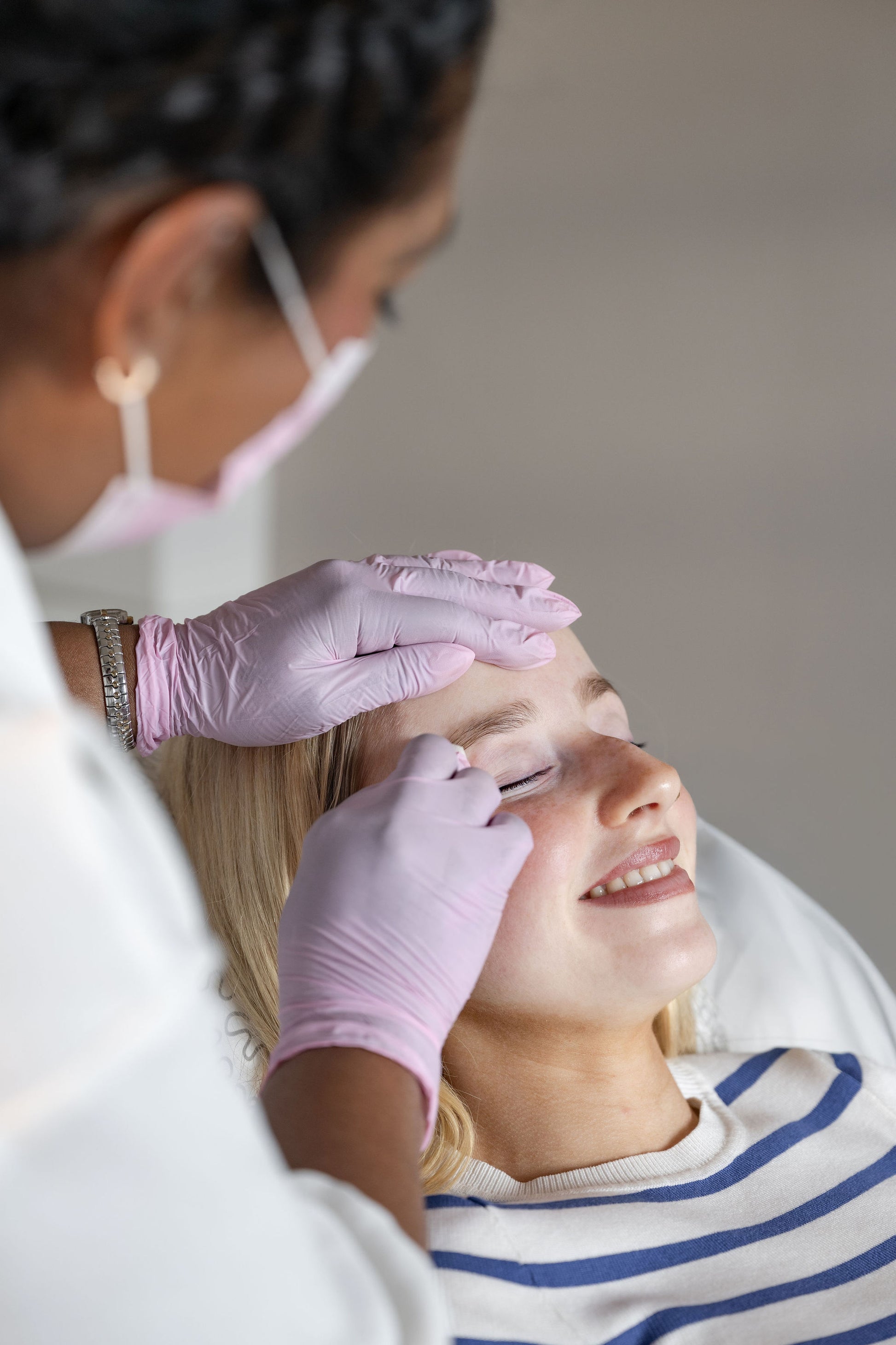 A close-up lifestyle shot of a beauty professional wearing pink gloves and a face mask, applying false eyelashes to a client's eye. The client is smiling and wearing a blue and white striped shirt, set against a soft, neutral background.
