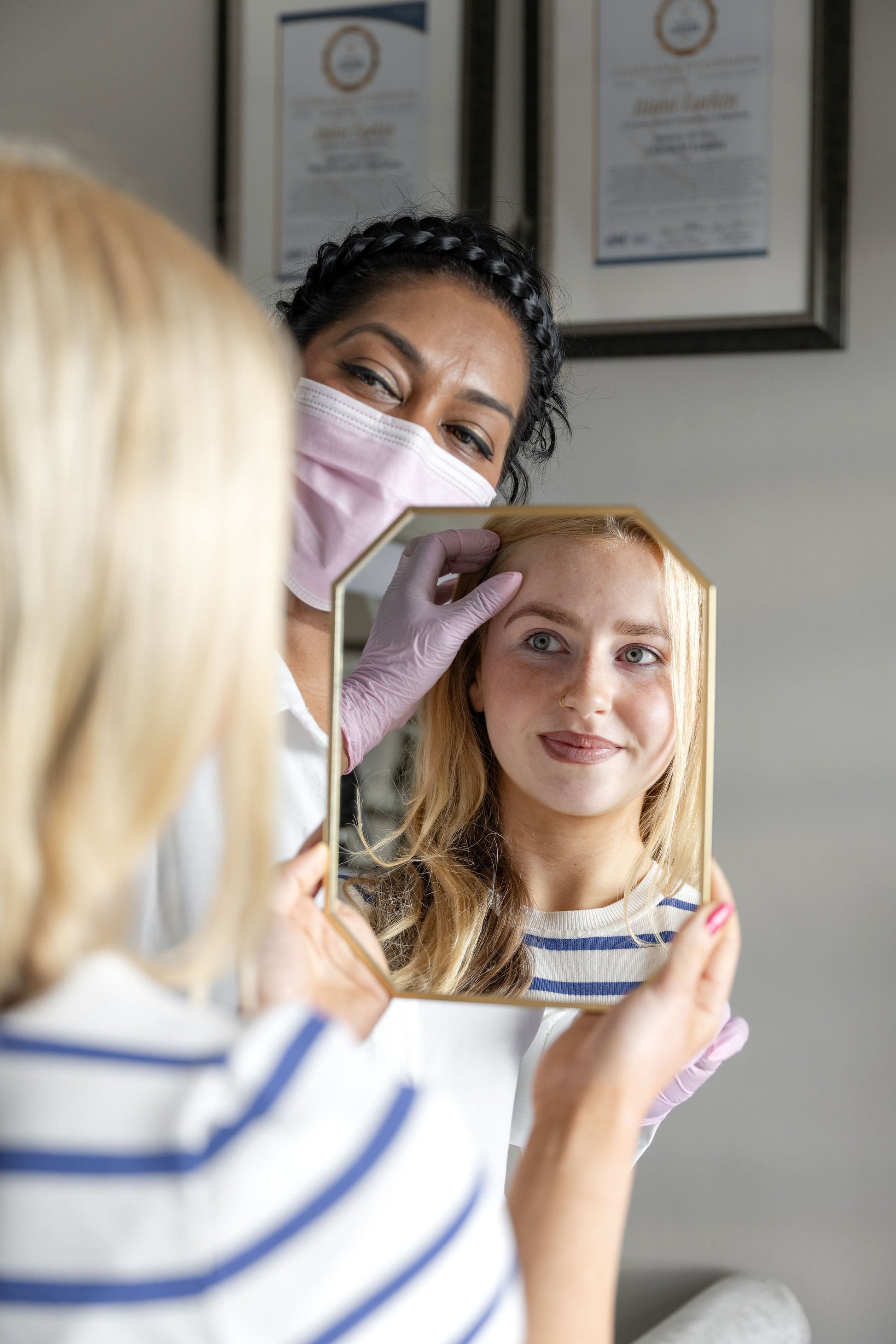 A lifestyle scene showing a beauty therapist wearing a pink face mask and purple gloves, using a small hexagonal mirror to examine a young girl's eyebrows and lashes. The girl is wearing a blue and white striped shirt and looking into the mirror, while the therapist stands behind her.