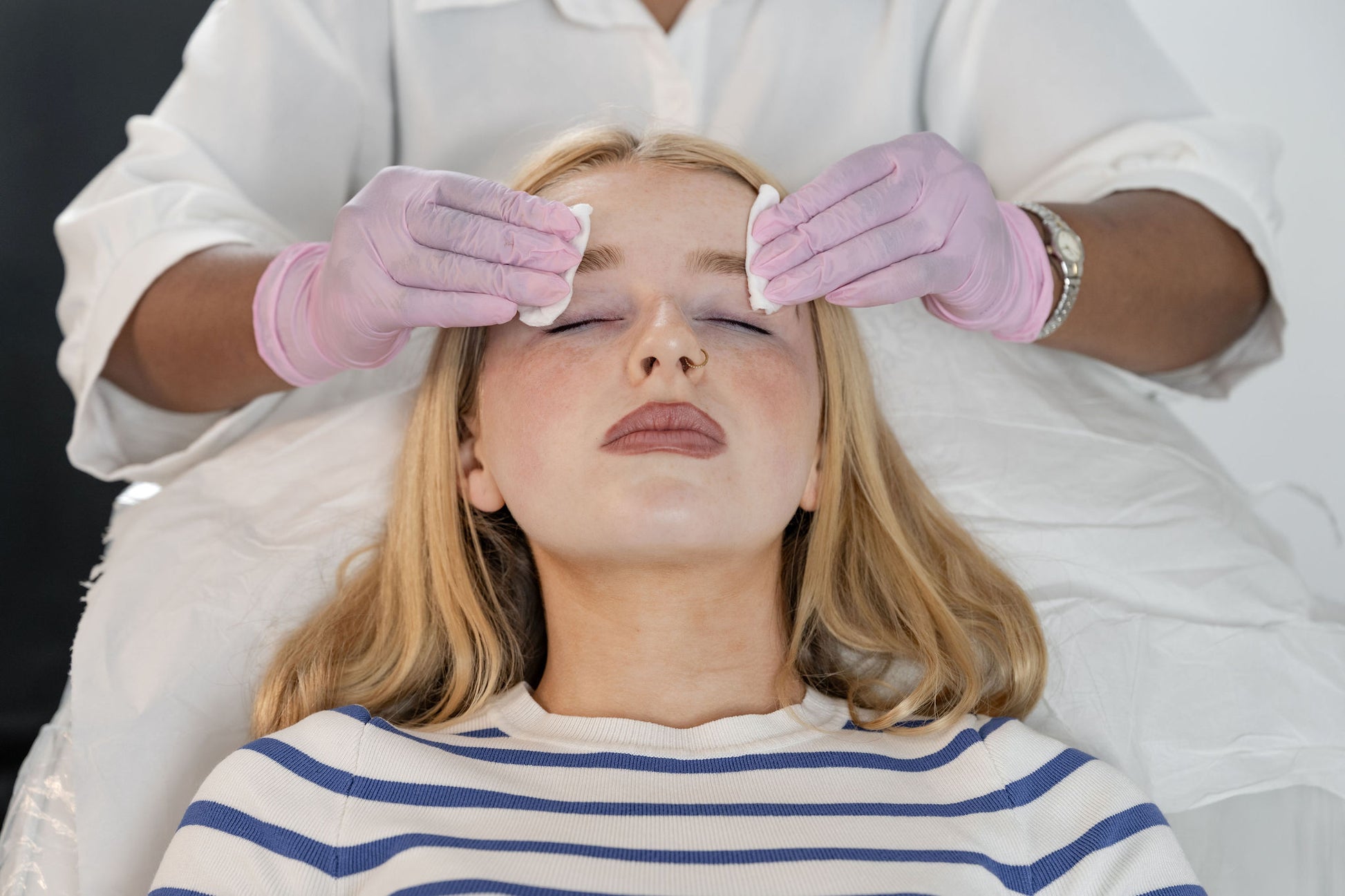 A woman with closed eyes lies on a treatment bed while another person in a white uniform and pink gloves wipes her face with cotton pads. She wears a blue and white striped shirt.
