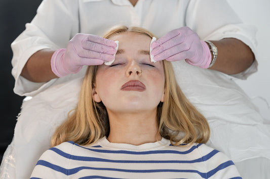 A woman with closed eyes lies on a treatment bed while another person in a white uniform and pink gloves wipes her face with cotton pads. She wears a blue and white striped shirt.