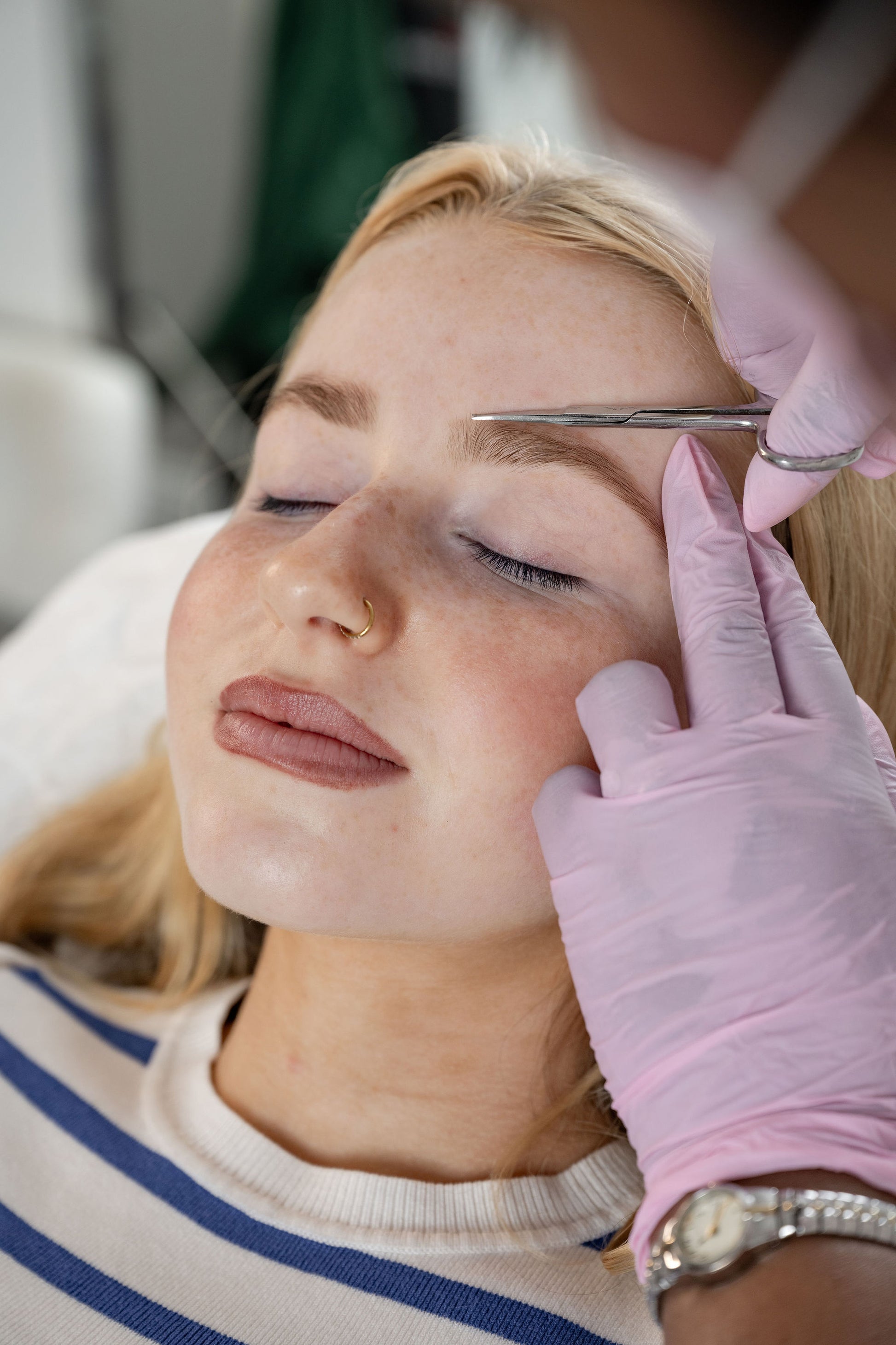 A close-up lifestyle shot of a woman receiving eyebrow grooming, with a manicured hand wearing a pink glove using small scissors on her eyebrow. The woman has blonde hair and is wearing a blue and white striped t shirt.