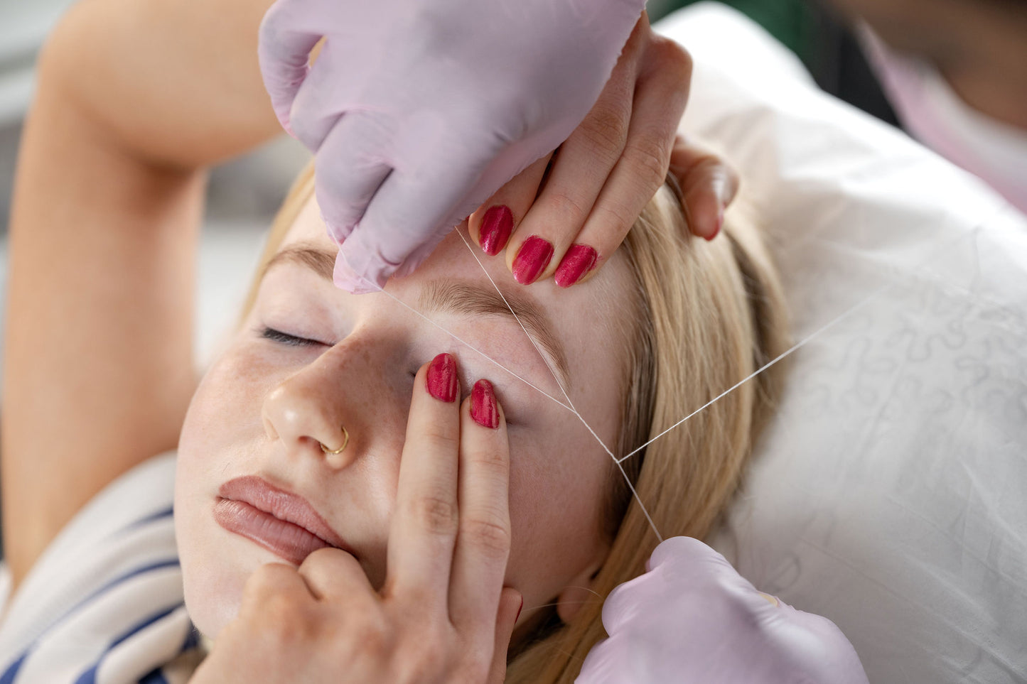 A close-up of a woman lying down with her eyes closed while a person wearing light purple gloves performs a facial thread lifting treatment. The therapist's hands with red nail polish hold the woman's face, and a fine thread is visible stretching across her forehead and eyebrow area.